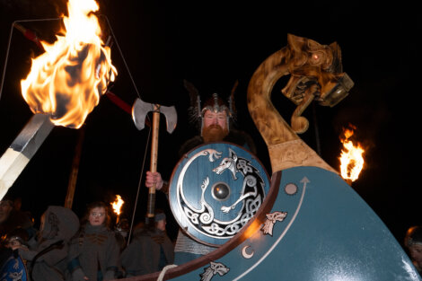 A person in Viking attire holds a shield and axe, standing next to a Viking ship replica with a dragon figurehead. Torches burn in the background.