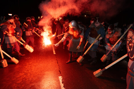 A group of people in Viking costumes holding torches and wearing helmets stand on a street at night, surrounded by smoke and firelight.
