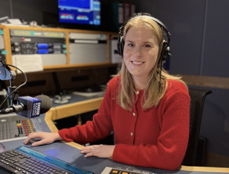 Person wearing headphones and a red sweater sits at a radio studio desk with equipment and a microphone.