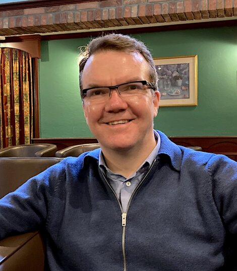 A person wearing glasses and a blue sweater sits in a room with green walls and framed artwork in the background. They are smiling.