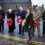 Six people stand solemnly holding wreaths of red poppies, participating in a remembrance ceremony on a wet street with stone buildings in the background.
