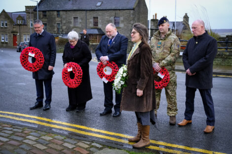 Six people stand solemnly holding wreaths of red poppies, participating in a remembrance ceremony on a wet street with stone buildings in the background.