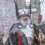 A man in Viking attire with a silver helmet, holding a shield and axe, stands in front of others in similar costumes. A building and a crowd are visible in the background.