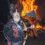 A person dressed in medieval armor with a horned helmet holds a large axe and a shield in front of a large bonfire at night.