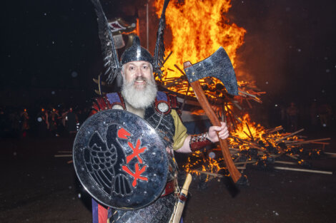 A person dressed in medieval armor with a horned helmet holds a large axe and a shield in front of a large bonfire at night.