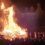 A large bonfire with a wooden structure burning intensely at night, surrounded by a crowd of people watching the event.