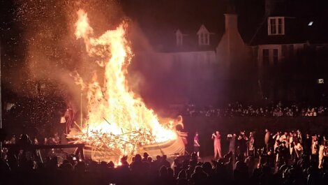 A large bonfire with a wooden structure burning intensely at night, surrounded by a crowd of people watching the event.