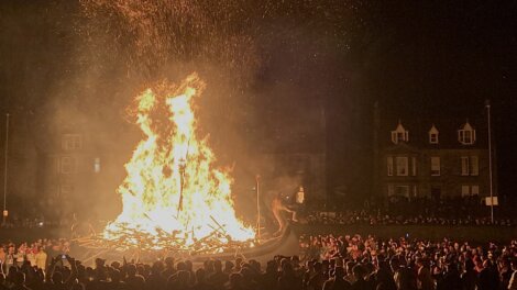 Large bonfire with a Viking-style boat burning amid a crowd of spectators at night. Embers and flames rise into the dark sky.