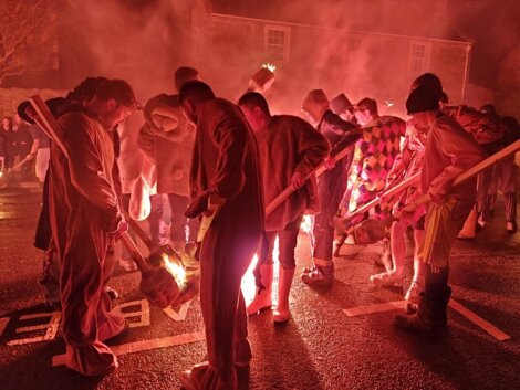 People in costumes gather around a small fire on a street at night, holding long poles. Smoke and warm light illuminate the scene.