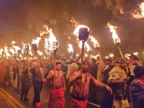 A group of people in costumes walk down a street at night, carrying large flaming torches on poles.