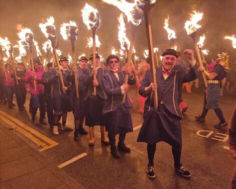 A group of people in matching outfits walk down a street at night, holding large flaming torches.