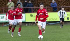 Soccer player in red jersey dribbles a ball on a field during a match, with teammates and spectators in the background.