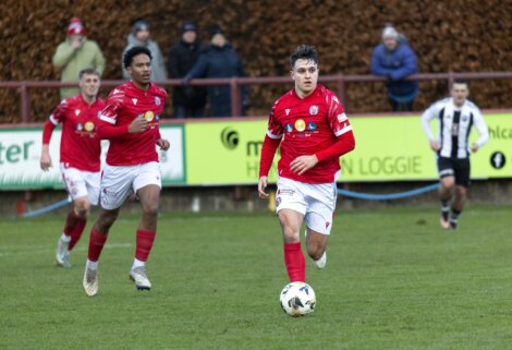 Soccer player in red jersey dribbles a ball on a field during a match, with teammates and spectators in the background.