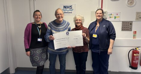 Four people stand indoors, holding a large presentation check for £10,000 addressed to Shetland Health Board. They are smiling and standing in front of an NHS Shetland sign.