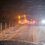 Two cars stopped on a snowy road at night, one slightly off the road near a fence, with headlights and taillights on, surrounded by falling snow.