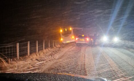 Two cars stopped on a snowy road at night, one slightly off the road near a fence, with headlights and taillights on, surrounded by falling snow.
