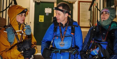 Three people wearing outdoor gear and headlamps talk indoors, preparing for an activity.