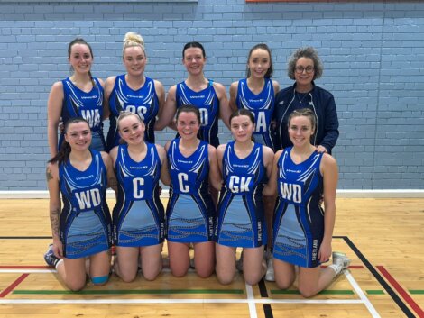 A netball team in blue uniforms poses for a group photo in a gymnasium.