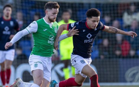 Two soccer players in action during a match, one wearing a green and white kit and the other in a dark blue kit with white shorts. Players are focused on the ball.