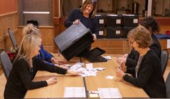 A woman empties a ballot box onto a table while four other people sort through the papers.