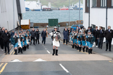 A group of people dressed in Viking-style costumes with shields walking down a street, followed by onlookers. There are buildings and a view of ships on the water in the background.
