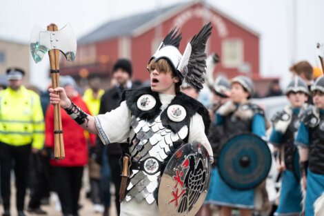 Person in Viking-inspired attire with a shield and axe leading a group in costume during an outdoor event.