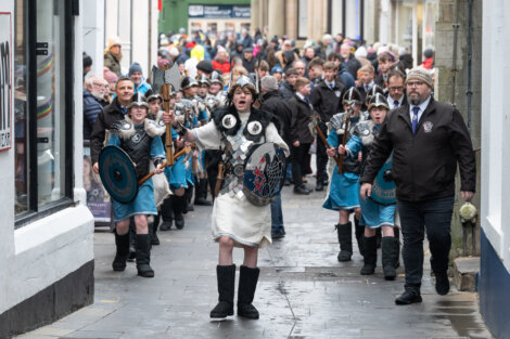 A group of people dressed as Vikings, some holding shields and axes, march through a narrow street, followed by a crowd.