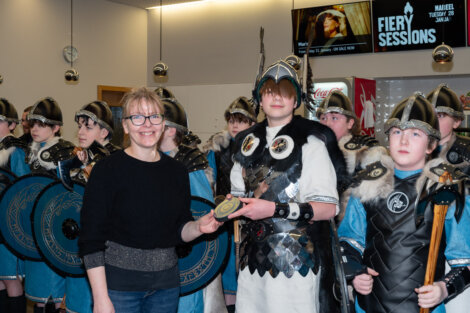 A woman presents an award to a young person dressed in Viking attire, surrounded by others similarly dressed, with shields and helmets. A screen in the background displays "Fiery Sessions".