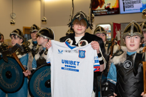A group of people in Viking costumes stands in the background. One person in the center holds up a signed football jersey while smiling.