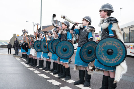 A row of young people in Viking costumes, holding shields, and blowing horns stand on a street. A group of adults is visible in the background.