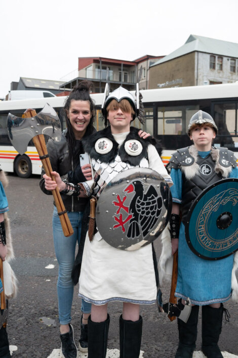 A woman smiles and stands with three children dressed in Viking costumes, including helmets and shields, in front of a bus.