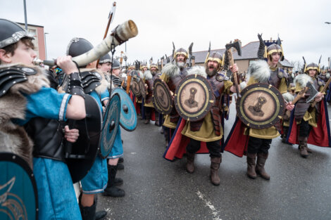 A group of people, dressed as Vikings with shields and helmets, march down a street. One person is blowing a large horn.