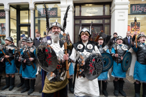A group of people in Viking costumes, with shields and helmets, stand in front of a shop named "Begg Shoes & Bags." The setting appears to be a street with onlookers in the background.