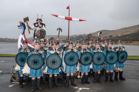 A group of people dressed as Vikings pose with shields and axes in front of a Viking ship model outdoors.