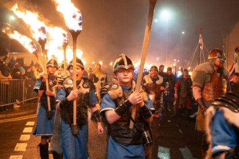 A group of people in Viking costumes march with flaming torches at night, surrounded by onlookers.