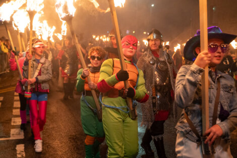 People in colorful costumes and helmets hold large torches while marching in a parade at night.