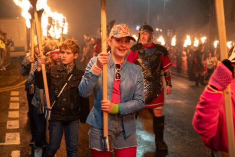 People marching with lit torches in a nighttime parade. A person in a blue jacket and colorful cap is in the foreground. More participants and onlookers are visible in the background.