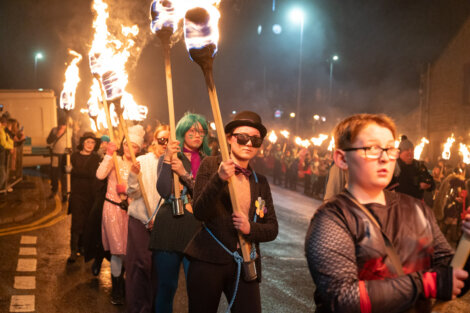A group of people in costumes carry flaming torches while walking down a street at night.