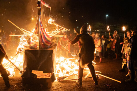 A group of people in Viking costumes stand around a burning wooden structure with a striped sail, holding torches during a nighttime event.