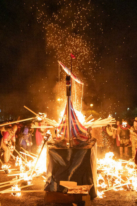 Viking ship replica set ablaze at night, surrounded by people dressed in costumes, sparks flying in the air.
