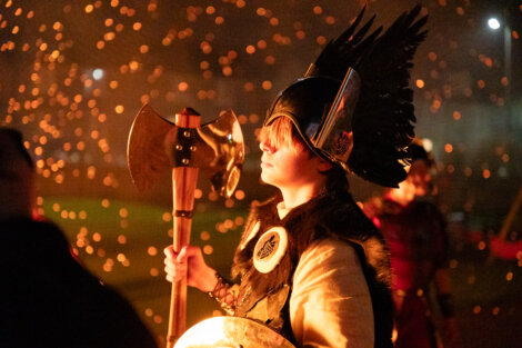 A person in Viking-style attire holds a large axe, surrounded by glowing embers.