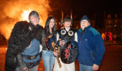 Four people pose in front of a large bonfire. One person wears Viking attire with a helmet and shield. The scene is set at night with others visible in the background.