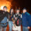 Four people pose in front of a large bonfire. One person wears Viking attire with a helmet and shield. The scene is set at night with others visible in the background.