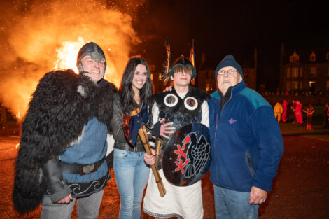 Four people pose in front of a large bonfire. One person wears Viking attire with a helmet and shield. The scene is set at night with others visible in the background.