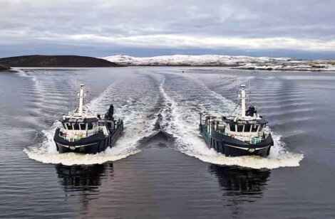Two boats navigate parallel paths on a calm body of water with snow-covered land in the background, leaving symmetrical wakes behind them.