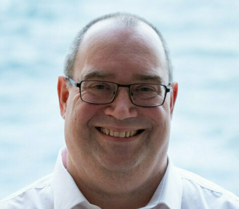 A man wearing glasses and a white shirt stands smiling in front of a blurry water background.