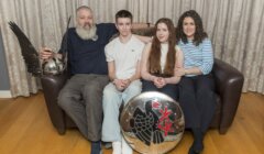 Family of four sitting on a couch; the father holds a helmet, the son wears a white shirt, the daughter holds a shield, and the mother is in a striped shirt. A curtain is in the background.