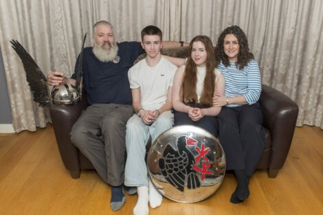 Family of four sitting on a couch; the father holds a helmet, the son wears a white shirt, the daughter holds a shield, and the mother is in a striped shirt. A curtain is in the background.