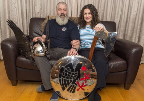 Man and woman sitting on a couch holding a Viking helmet, shield with a raven design, and an axe.