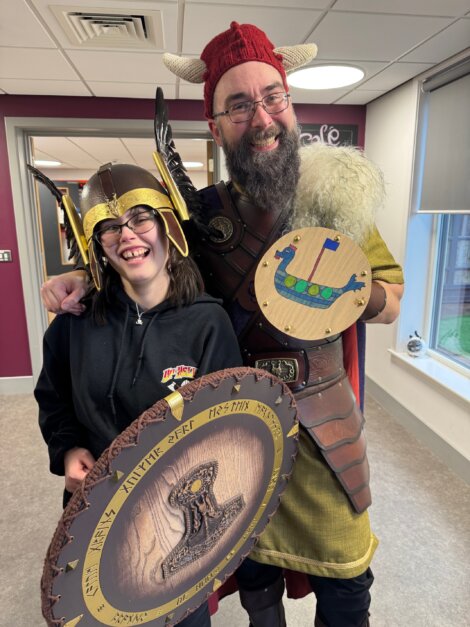Two people dressed in Viking costumes, one with a horned helmet and the other with a winged helmet, holding round shields with designs. They are smiling in an indoor setting.